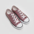 A pair of low-top canvas sneakers in a dusty rose or mauve pink color, featuring white laces, white rubber toe caps, and black trim, displayed on a white brick background.