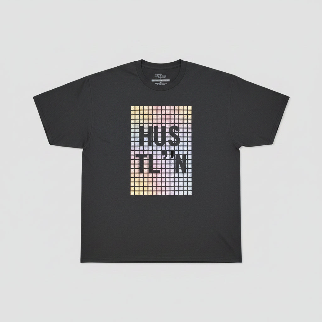 A black short-sleeve t-shirt with a large pixelated graphic on the front reading 'HUSTLE' in a gold, orange, and pink gradient, hanging against a light wood paneled wall.