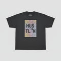 A black short-sleeve t-shirt with a large pixelated graphic on the front reading 'HUSTLE' in a gold, orange, and pink gradient, hanging against a light wood paneled wall.